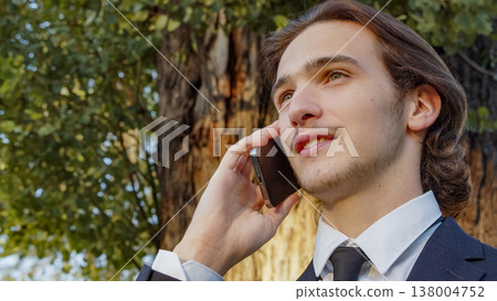 Portrait of the Businessman in a business suit in the park with mobile phone. Smiling businessman talking on a cell phone., outdoors. Young man in a business suit talking on a smartphone in nature Portrait of the Businessman in a business suit in the park with mobile phone. Smiling businessman talking on a cell phone., outdoors. Young man in a business suit talking on a smartphone in nature 138004752