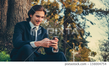 Happy man in a business suit sets outdoors and using a smartphone. Portrait of the young man in a business suit with mobile phone, in the park. Smiling businessman in city looking to a cell phone. Happy man in a business suit sets outdoors and using a smartphone. Portrait of the young man in a business suit with mobile phone, in the park. Smiling businessman in city looking to a cell phone. 138004754