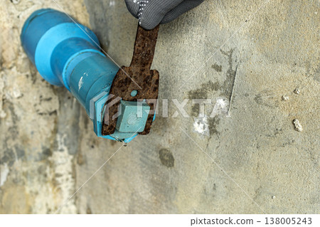 Close-Up of Gloved Worker Hand Gripping a Heavily Corroded Rusty Metal Wrench Turning Blue PVC Plastic Water Pipe Valve Fitting on Rough Concrete Floor 138005243
