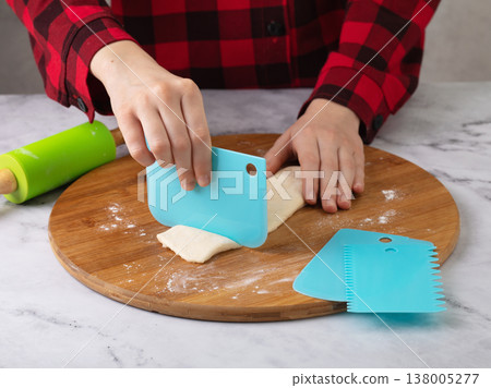 Child's hands using blue scraper to cut dough into pieces on wooden cutting board surrounded by rolling pin and other scrapers Child's hands using blue scraper to cut dough into pieces on wooden cutting board surrounded by rolling pin and other scrapers 138005277