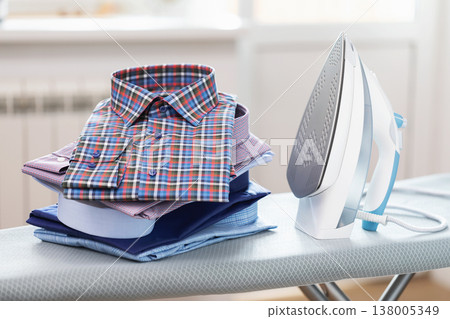 Folded long sleeve shirts and iron on ironing board in room Folded long sleeve shirts and iron on ironing board in room 138005349