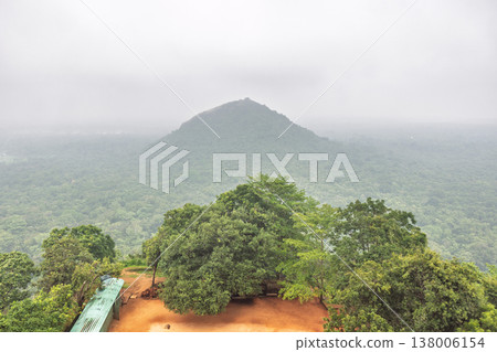 Sigiriya ancient rock fortress in Sri Lanka. Panoramic view of a lush green tropical forest and distant mountain under an overcast, hazy sky, serene landscape Sigiriya ancient rock fortress in Sri Lanka. Panoramic view of a lush green tropical forest and distant mountain under an overcast, hazy sky, serene landscape 138006154
