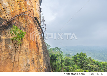 Sigiriya ancient rock fortress in Sri Lanka. Ancient Rock Fortress Wall with Unique Railings and Lush Forest Vista Under a Hazy Sky, Tropical Landscape Sigiriya ancient rock fortress in Sri Lanka. Ancient Rock Fortress Wall with Unique Railings and Lush Forest Vista Under a Hazy Sky, Tropical Landscape 138006169