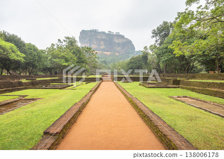 Sigiriya ancient rock fortress with gardens in Sri Lanka. Ancient Pathway Leading to Majestic Rock Formation Amidst Lush Green Gardens and Historical Ruins on a Cloudy Day Sigiriya ancient rock fortress with gardens in Sri Lanka. Ancient Pathway Leading to Majestic Rock Formation Amidst Lush Green Gardens and Historical Ruins on a Cloudy Day 138006178