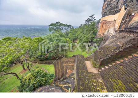 Sigiriya ancient rock fortress in Sri Lanka. Ancient Staircase Carved into a Massive Rock Formation Overlooking Lush Green Forest and Distant Horizon Under Cloudy Sky Sigiriya ancient rock fortress in Sri Lanka. Ancient Staircase Carved into a Massive Rock Formation Overlooking Lush Green Forest and Distant Horizon Under Cloudy Sky 138006193