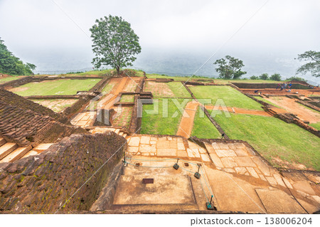Sigiriya ancient rock fortress in Sri Lanka. Ancient architectural ruins with lush green terraces and a solitary tree under an overcast sky, showcasing historical landscape 138006204