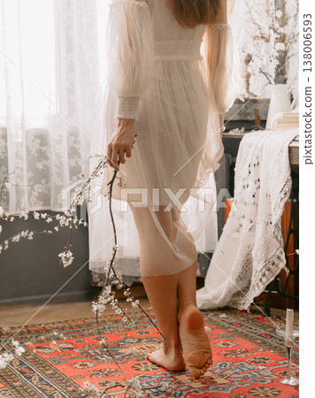 Barefoot woman in sheer dress holding blossom branch in bright airy interior. Barefoot woman in sheer dress holding blossom branch in bright airy interior. 138006593