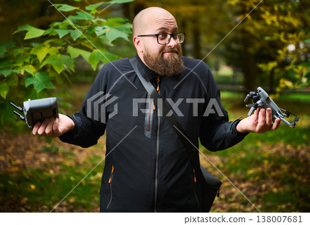 Man shrugs while holding drone and controller in hands. Bearded man looks confused in park comparing devices, unsure what to do, concept of choice, uncertainty and technology Man shrugs while holding drone and controller in hands. Bearded man looks confused in park comparing devices, unsure what to do, concept of choice, uncertainty and technology 138007681