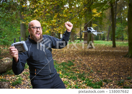 Man celebrates while flying a drone with remote controller. Bearded man operating quadcopter in a park, reacting with excitement during flight, concept of modern technology and hobby Man celebrates while flying a drone with remote controller. Bearded man operating quadcopter in a park, reacting with excitement during flight, concept of modern technology and hobby 138007682
