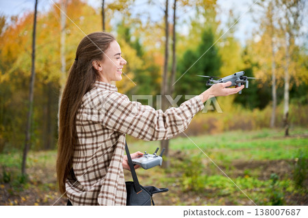 Woman launches drone from her hand while smiling and holding controller. Side view of young woman in autumn park starting quadcopter flight, concept of hobby, technology and outdoor activity 138007687