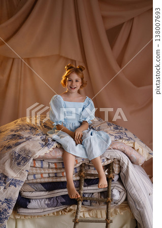 Little girl in blue dress sitting on stacked mattresses like princess and the pea, studio drapery background 138007693
