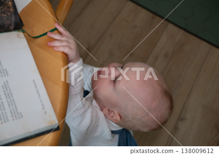 Baby reaches for book on table in home interior with soft light, early exploration and interaction with object in everyday domestic setting. Child development, parenting content, learning activity Baby reaches for book on table in home interior with soft light, early exploration and interaction with object in everyday domestic setting. Child development, parenting content, learning activity 138009612