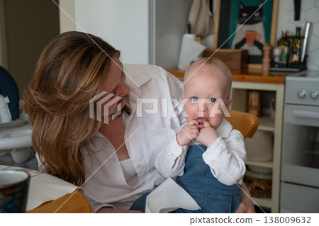 Mother holds baby in kitchen interior with natural light while baby holds tissue, everyday childcare moment and interaction in home setting. Family bonding, parenting, child care routine, lifestyle Mother holds baby in kitchen interior with natural light while baby holds tissue, everyday childcare moment and interaction in home setting. Family bonding, parenting, child care routine, lifestyle 138009632