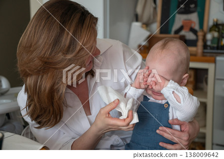 Mother wipes baby nose with tissue in kitchen interior with natural light and home setting, everyday childcare routine and hygiene care moment. Childcare routine, parenting content, family Mother wipes baby nose with tissue in kitchen interior with natural light and home setting, everyday childcare routine and hygiene care moment. Childcare routine, parenting content, family 138009642