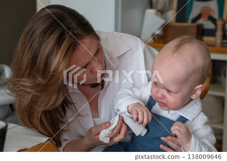 Mother wipes baby nose with tissue in kitchen interior with natural light and home setting, everyday care moment and parenting routine in domestic environment. Childcare routine, parenting, family Mother wipes baby nose with tissue in kitchen interior with natural light and home setting, everyday care moment and parenting routine in domestic environment. Childcare routine, parenting, family 138009646