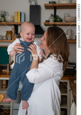 Mother holds smiling baby in kitchen interior with natural light and home decor, joyful interaction and everyday family moment in domestic setting. Family bonding, motherhood, parenting Mother holds smiling baby in kitchen interior with natural light and home decor, joyful interaction and everyday family moment in domestic setting. Family bonding, motherhood, parenting 138009655