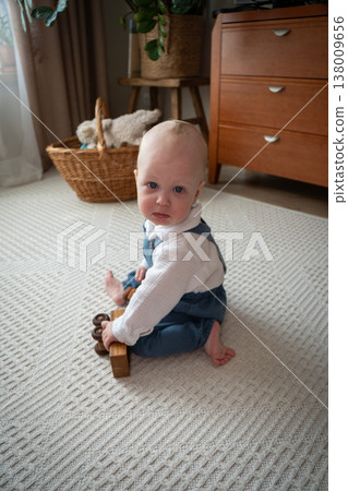 Baby sits on carpet holding wooden toy in home interior with natural light, cozy setting, early development and play activity in everyday domestic environment. Child development, parenting content Baby sits on carpet holding wooden toy in home interior with natural light, cozy setting, early development and play activity in everyday domestic environment. Child development, parenting content 138009656
