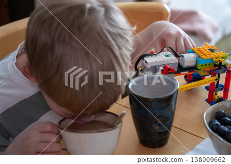 Boy drinks from cup at table with construction toy in home interior with natural light, everyday meal moment and activity in domestic setting. Childhood activity, family life, daily routine, lifestyle Boy drinks from cup at table with construction toy in home interior with natural light, everyday meal moment and activity in domestic setting. Childhood activity, family life, daily routine, lifestyle 138009662