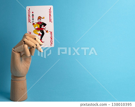 Wooden hand holds a joker playing card against a blue background in a simple setup Wooden hand holds a joker playing card against a blue background in a simple setup 138010395