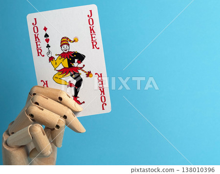 Wooden hand holding a joker playing card in front of a blue background indoors Wooden hand holding a joker playing card in front of a blue background indoors 138010396