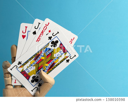 Playing cards held by a wooden hand on a blue background with different card ranks shown and joker present in the hand Playing cards held by a wooden hand on a blue background with different card ranks shown and joker present in the hand 138010398