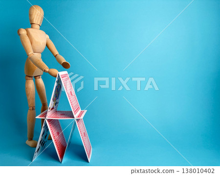 Wooden figure builds a house of cards on a blue background during daytime in a simple setting Wooden figure builds a house of cards on a blue background during daytime in a simple setting 138010402