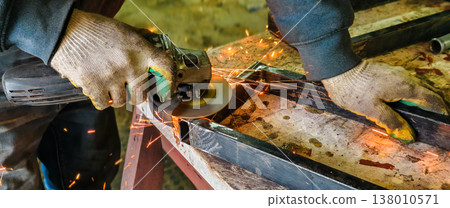 Worker wearing protective gloves safely operating an angle grinder to cut a metal profile, creating a shower of bright orange sparks against a blurred industrial workshop background. 138010571
