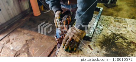 Worker in safety gloves grinding a steel frame with an angle grinder, sending bright sparks across a busy industrial workshop during metal fabrication and repair. Worker in safety gloves grinding a steel frame with an angle grinder, sending bright sparks across a busy industrial workshop during metal fabrication and repair. 138010572