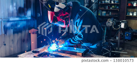 Skilled arc welder wearing a protective mask and gloves, joining metal components with bright blue light and sparks in a workshop environment, demonstrating craftsmanship and industrial safety. Skilled arc welder wearing a protective mask and gloves, joining metal components with bright blue light and sparks in a workshop environment, demonstrating craftsmanship and industrial safety. 138010573