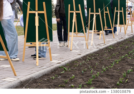 Row of wooden easels with green boards on a paved walkway in a park. People standing behind blank canvases during an outdoor art class or public exhibition. Spring plants emerging in foreground Row of wooden easels with green boards on a paved walkway in a park. People standing behind blank canvases during an outdoor art class or public exhibition. Spring plants emerging in foreground 138010876