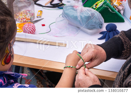 The grandmother's older hands tie a handmade green thread bracelet on the child's wrist. Representatives of different generations are engaged in needlework at a table in a workshop with beads and The grandmother's older hands tie a handmade green thread bracelet on the child's wrist. Representatives of different generations are engaged in needlework at a table in a workshop with beads and 138010877