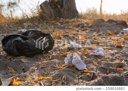 Black plastic garbage bag and discarded tissues on sandy ground with dry autumn leaves. Environmental pollution and littering in nature concept. Human impact on ecology Black plastic garbage bag and discarded tissues on sandy ground with dry autumn leaves. Environmental pollution and littering in nature concept. Human impact on ecology 138010881