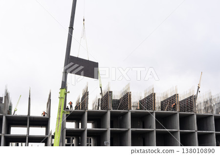 Green crane lifting a heavy dark panel at a construction site. Multi-story concrete building framework with workers in safety vests. Urban development and industrial engineering concept. Copy space Green crane lifting a heavy dark panel at a construction site. Multi-story concrete building framework with workers in safety vests. Urban development and industrial engineering concept. Copy space 138010890