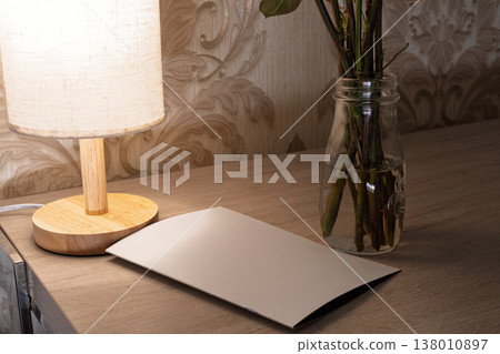 Blank folded card mockup on a wooden table next to a glowing lamp and a glass vase with plant stems. Warm interior lighting with floral wallpaper background. Copy space for invitation design Blank folded card mockup on a wooden table next to a glowing lamp and a glass vase with plant stems. Warm interior lighting with floral wallpaper background. Copy space for invitation design 138010897
