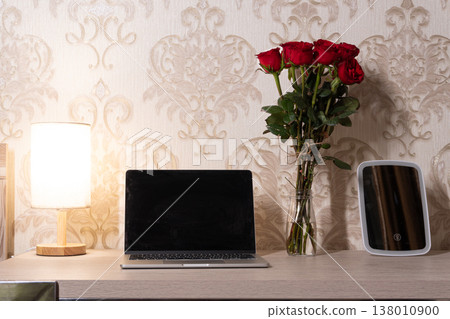 Home office workspace with open laptop and blank screen. Red roses in a glass vase on a wooden desk next to a warm table lamp and makeup mirror. Elegant damask wallpaper background mockup Home office workspace with open laptop and blank screen. Red roses in a glass vase on a wooden desk next to a warm table lamp and makeup mirror. Elegant damask wallpaper background mockup 138010900