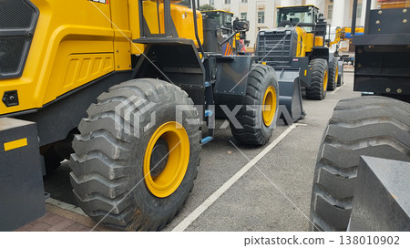 Close-up of heavy yellow wheel loaders parked in a row. Large construction machinery with massive black tires and front buckets. Industrial earthmoving equipment on asphalt 138010902