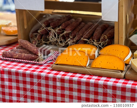 Cheese wheels and sausage are displayed on a market counter with a checkered tablecloth. Traditional food, artisanal production, local flavor, and the inviting richness of deli commerce. Cheese wheels and sausage are displayed on a market counter with a checkered tablecloth. Traditional food, artisanal production, local flavor, and the inviting richness of deli commerce. 138010937