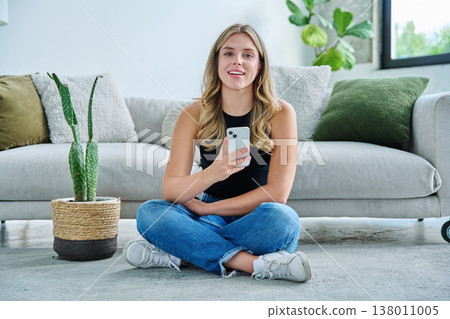 Smiling young woman with smartphone looking at camera sitting at home 138011005