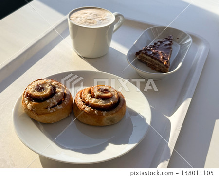Coffee cup with foam and pastries with cake slice on tray in sunlight. Relaxation moment, cafe lifestyle, leisure break, enjoyment, calm atmosphere, everyday pleasure 138011105