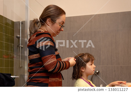 Mother braids her daughter's hair in a bathroom during a calm morning family routine. 138011227