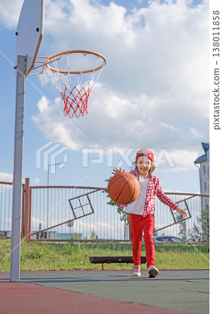 A happy little laughing girl in a baseball cap leads a basketball on the playground of a kindergarten 138011858