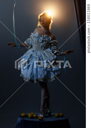 Young woman in Rococo dress holding ropes on small pedestal with lemons under bright light Young woman in Rococo dress holding ropes on small pedestal with lemons under bright light 138011869