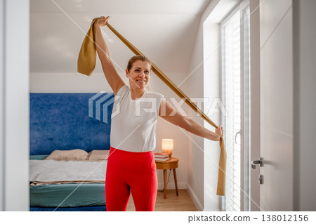 Smiling middle aged woman exercising with resistance band at home. Smiling middle aged woman exercising with resistance band at home. 138012156