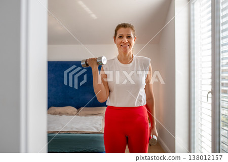Smiling middle aged woman exercising with dumbbells at home. 138012157
