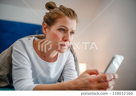 Middle aged woman scrolling on smartphone in bed. Middle aged woman scrolling on smartphone in bed. 138012160