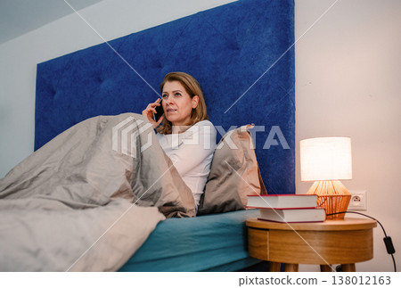 Mature woman making phone call while sitting in bed at home. Mature woman making phone call while sitting in bed at home. 138012163