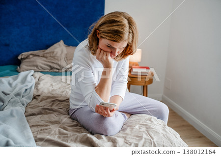 Mature woman using smartphone while sitting on bed. Mature woman using smartphone while sitting on bed. 138012164