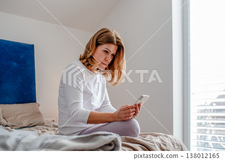 Mature woman using smartphone while sitting on bed. Mature woman using smartphone while sitting on bed. 138012165