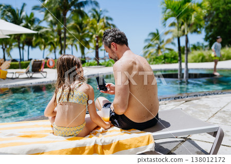 Family relaxing by tropical resort pool, child with diabetes wears glucose monitor. 138012172
