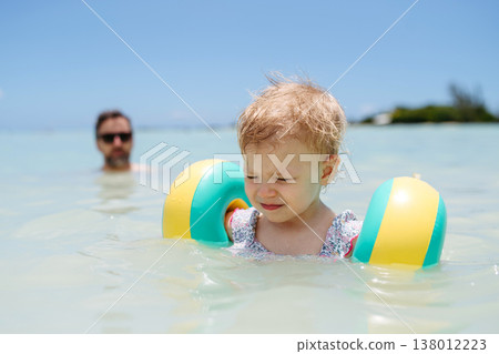 Toddler girl swimming in sea with arm floaties while father in background. 138012223